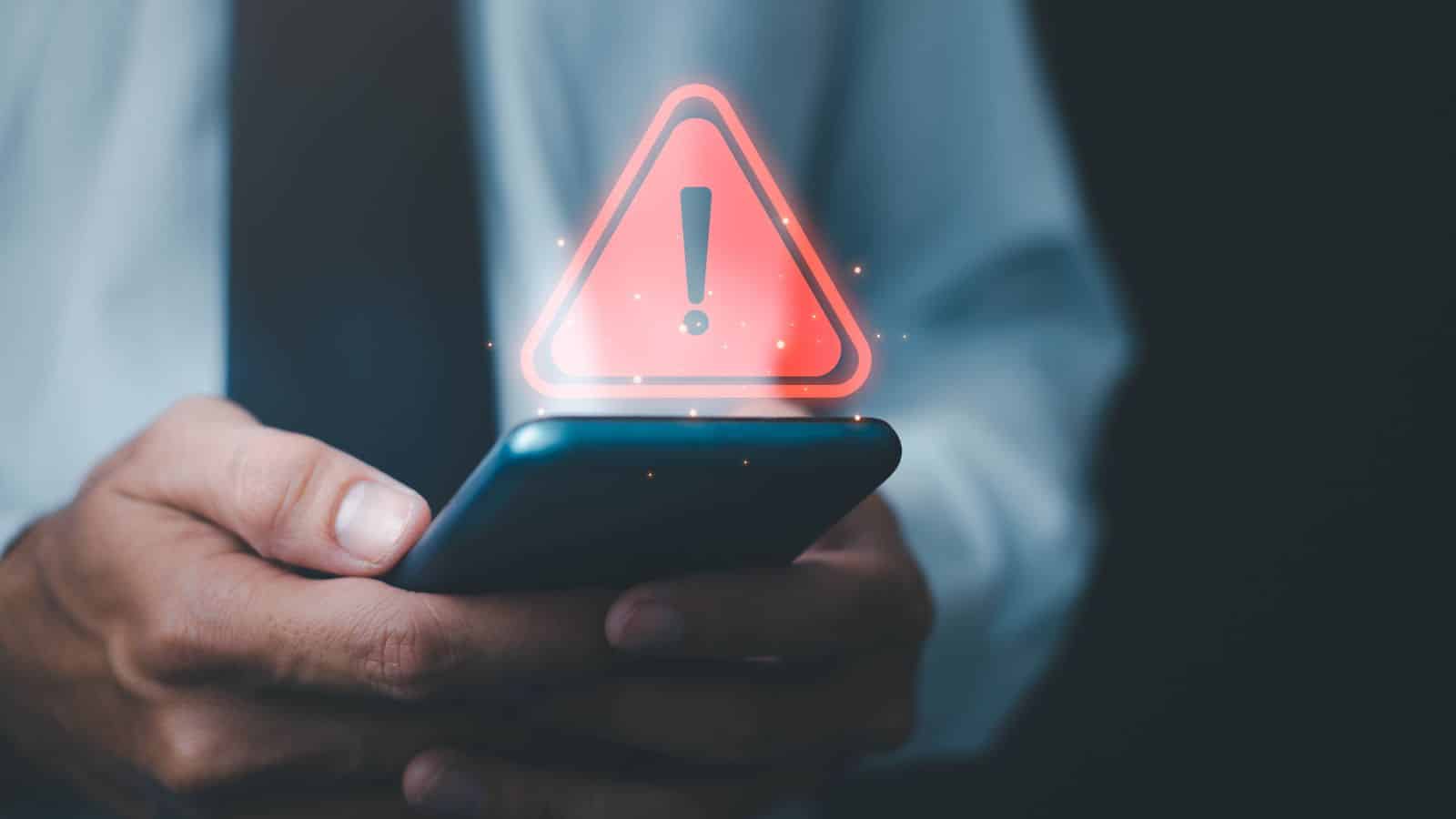 A person in business attire holds a smartphone with both hands. A red warning triangle with an exclamation mark appears above the phone, hinting at a vacation scam alert or a travel protection notification for travel credit cards.