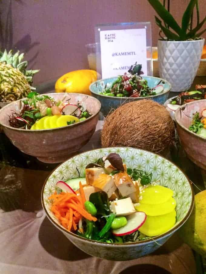 Four ceramic bowls filled with colorful salads containing various vegetables and tofu sit on a table. A coconut, pineapple, mango, and potted plant are also displayed around the bowls as decoration.