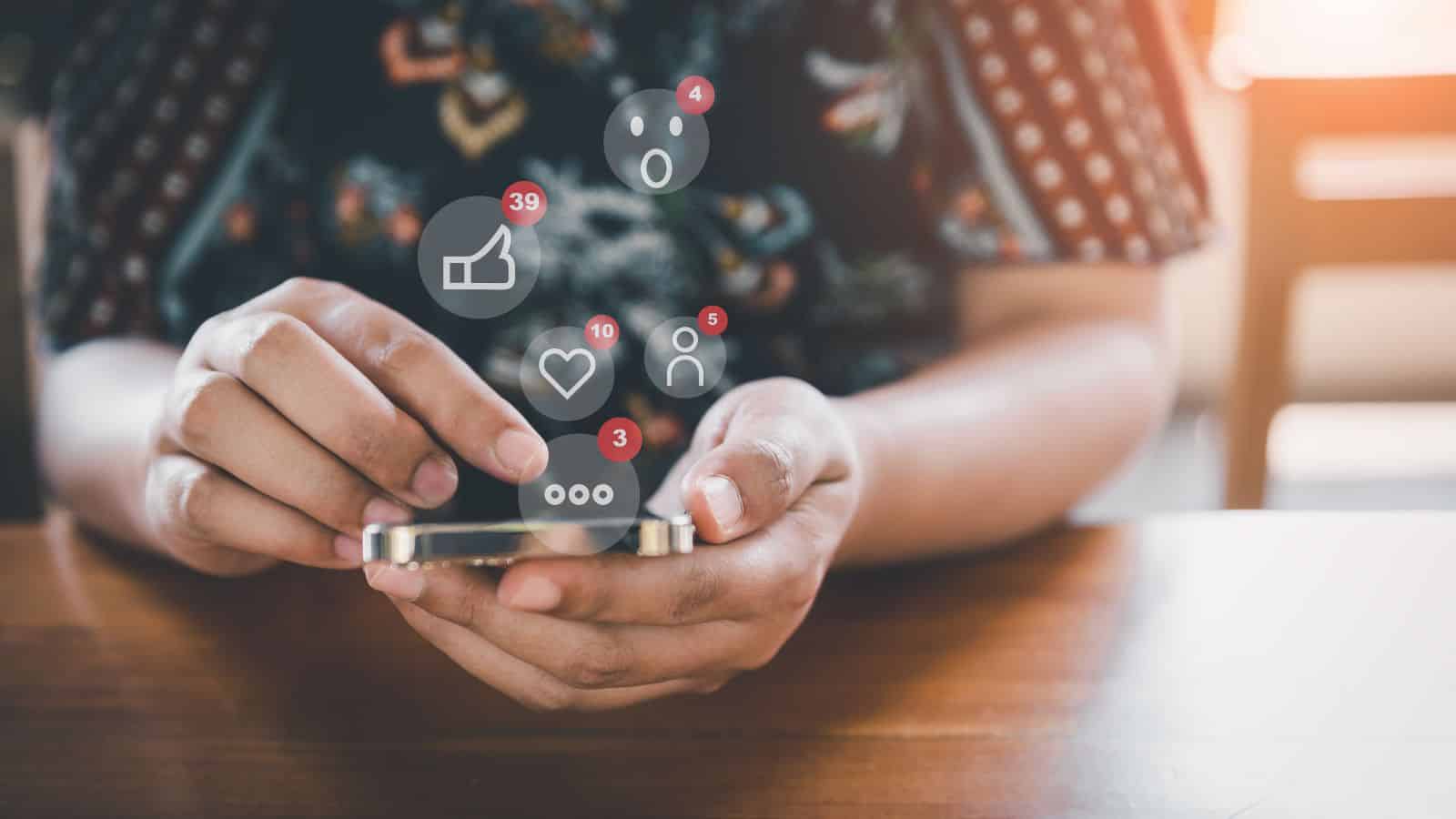 A person holding a smartphone sits at a wooden table, with social media notification icons for likes, comments, and messages digitally superimposed above the device.