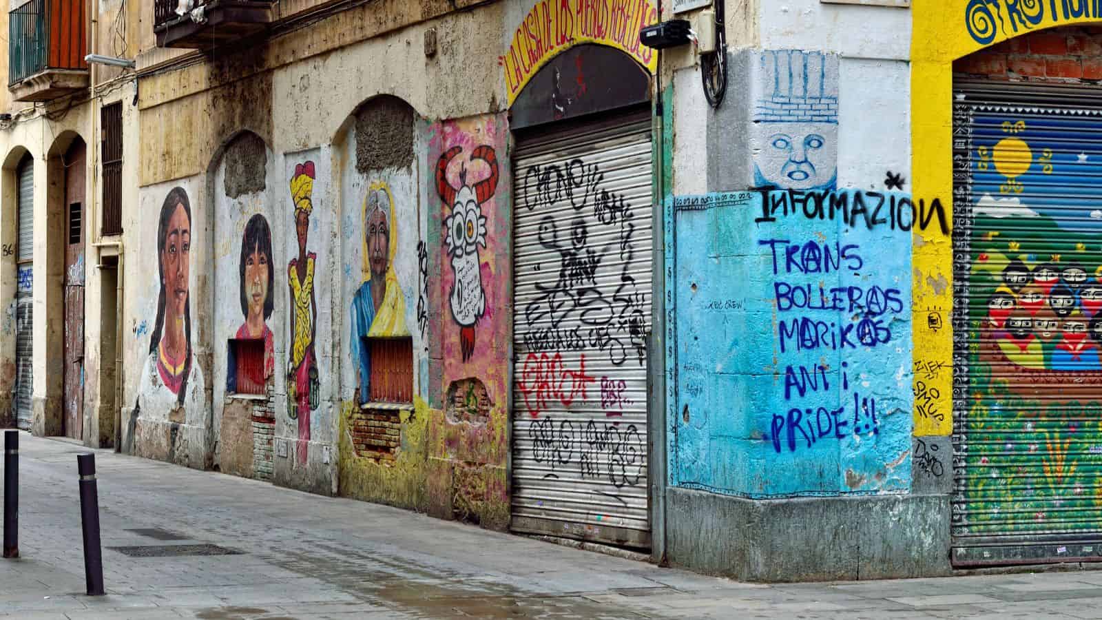A row of urban buildings with graffiti and murals on metal shutters and walls, including portraits, cartoon-style faces, and blue spray-painted text about trans, Bollebas, Morikos, anti-pride, and information.