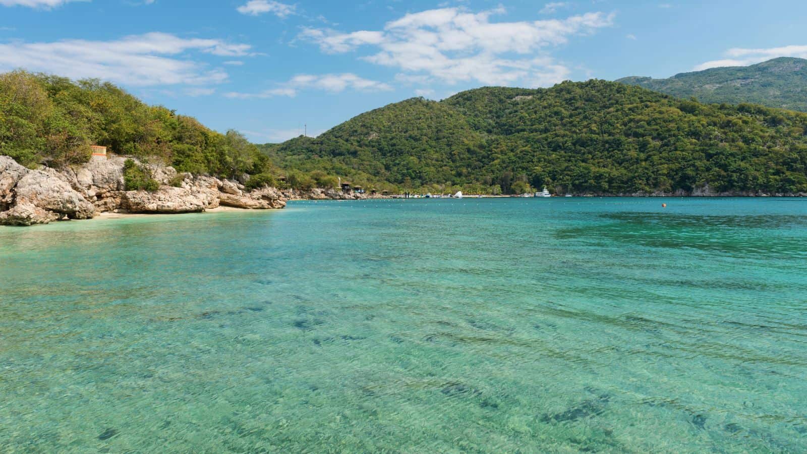 Clear turquoise water with gentle ripples in the foreground, rocky shoreline and green hills covered with trees in the background, under a partly cloudy sky.