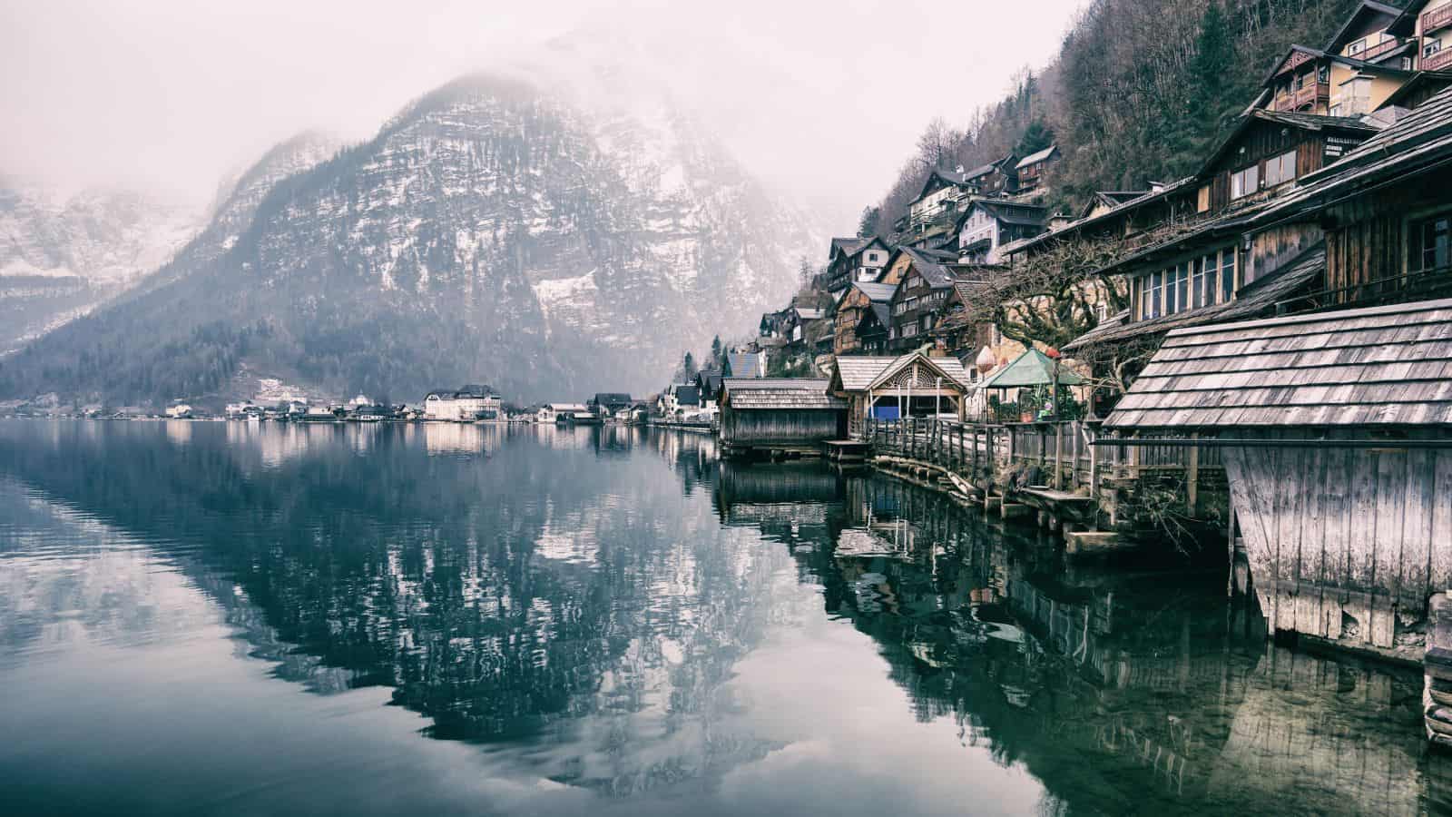 A lakeside village with wooden houses lines the shore, with forested mountains in the background partially covered in snow and mist. The calm water reflects the buildings and mountains, creating a mirror-like effect.