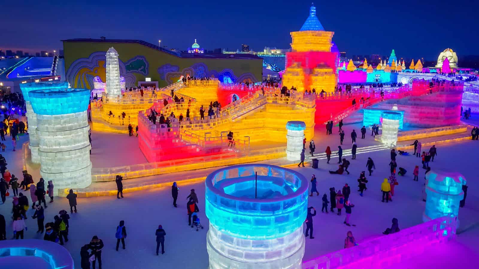 People walk among large illuminated ice sculptures and castles at night; the structures glow in various colors including blue, yellow, pink, and orange, with more lights and artworks visible in the background.
