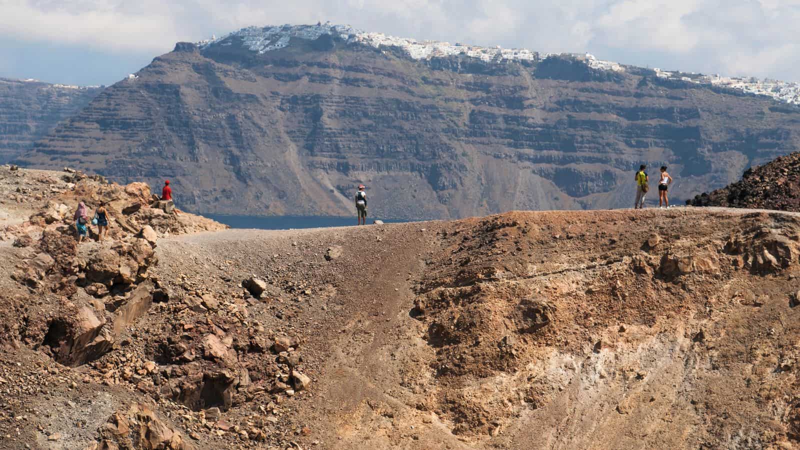 People stand and walk on a rocky path with uneven terrain. In the background, there is a steep, rugged mountain with white buildings along its ridge under a partly cloudy sky.