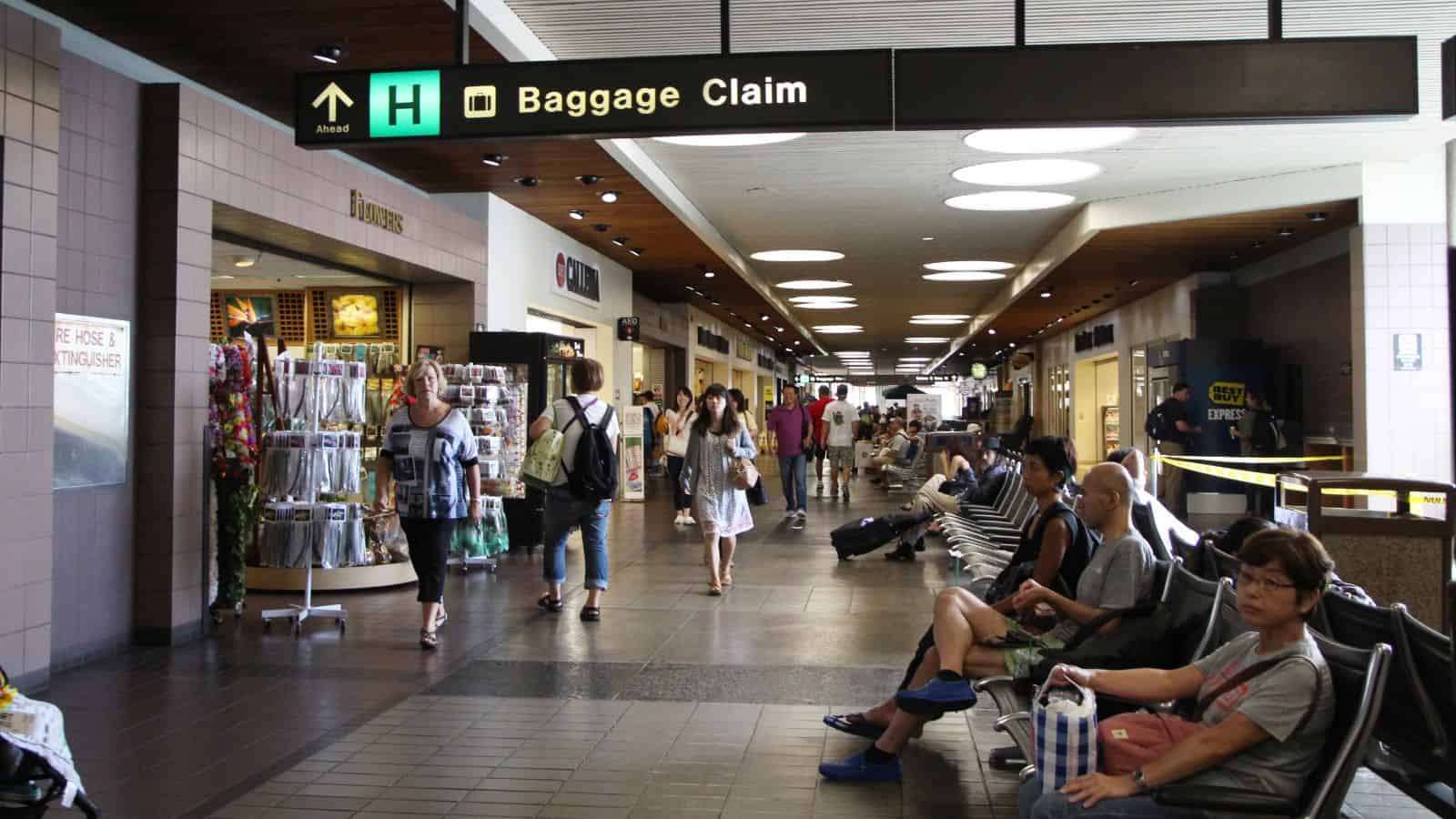 People walk and sit in an airport terminal with shops on the left and a sign overhead indicating directions to baggage claim. Some travelers carry bags, while others are seated and waiting.