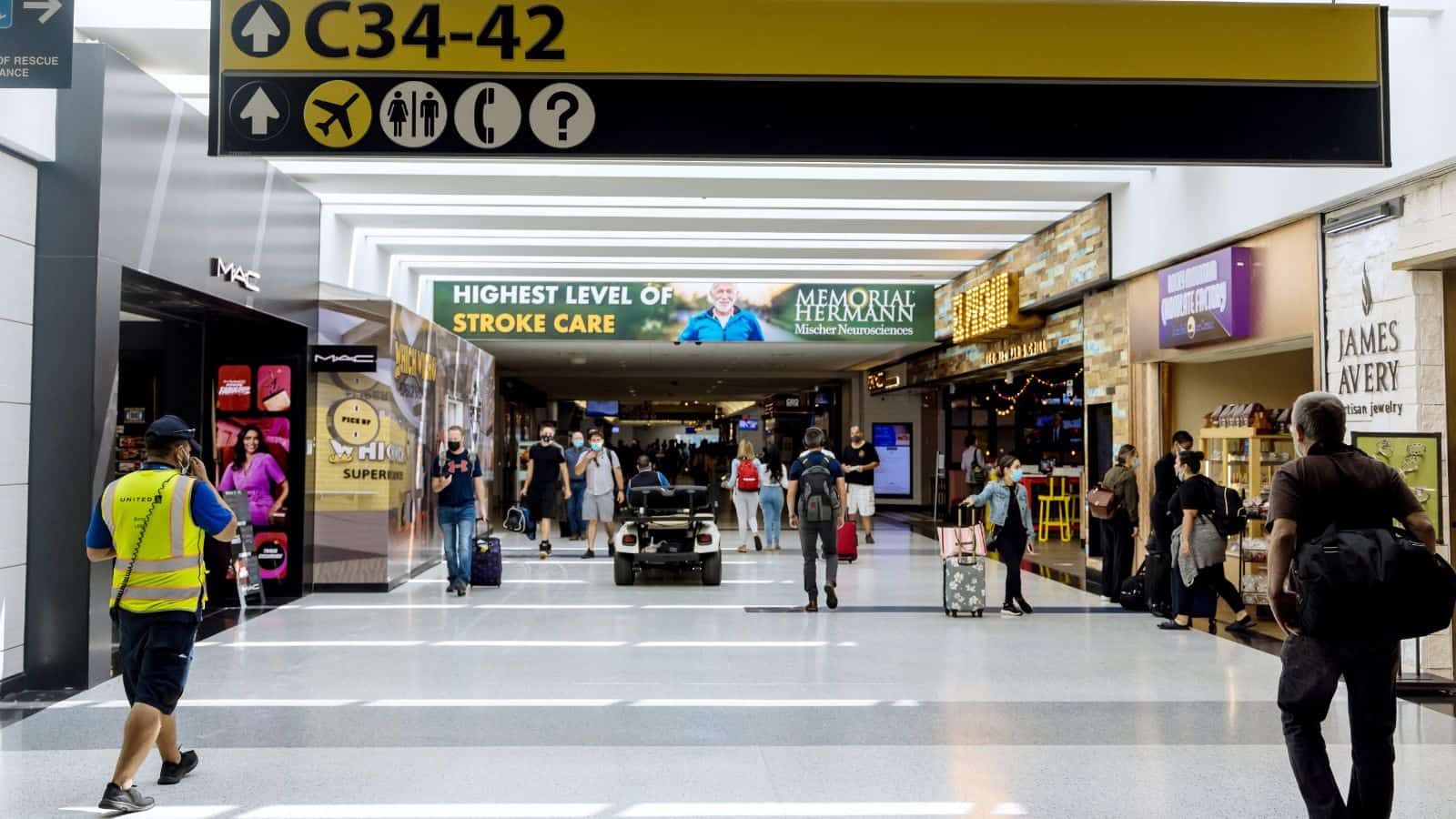 Travelers walk through an airport terminal with signs for gates C34-42 overhead. Various shops and restaurants line the sides, and a few people are pulling suitcases. A cart with passengers is in the center.