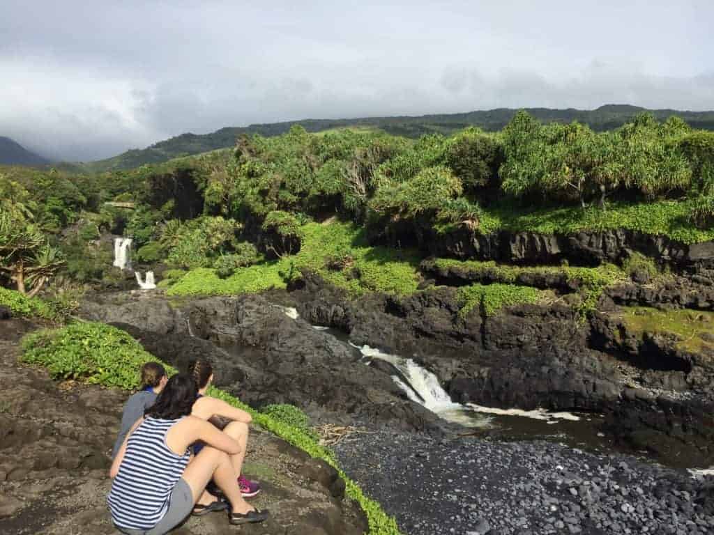 Three people sit on a rocky ledge overlooking a lush, green landscape with several small waterfalls and streams flowing through black volcanic rock under a cloudy sky.