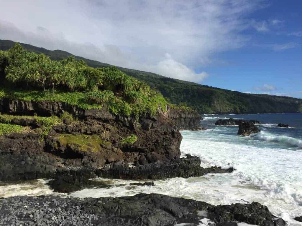 Rocky coastline with green vegetation on cliffs overlooking the ocean, waves crashing onto dark rocks below, and hills covered in greenery in the background under a partly cloudy sky.