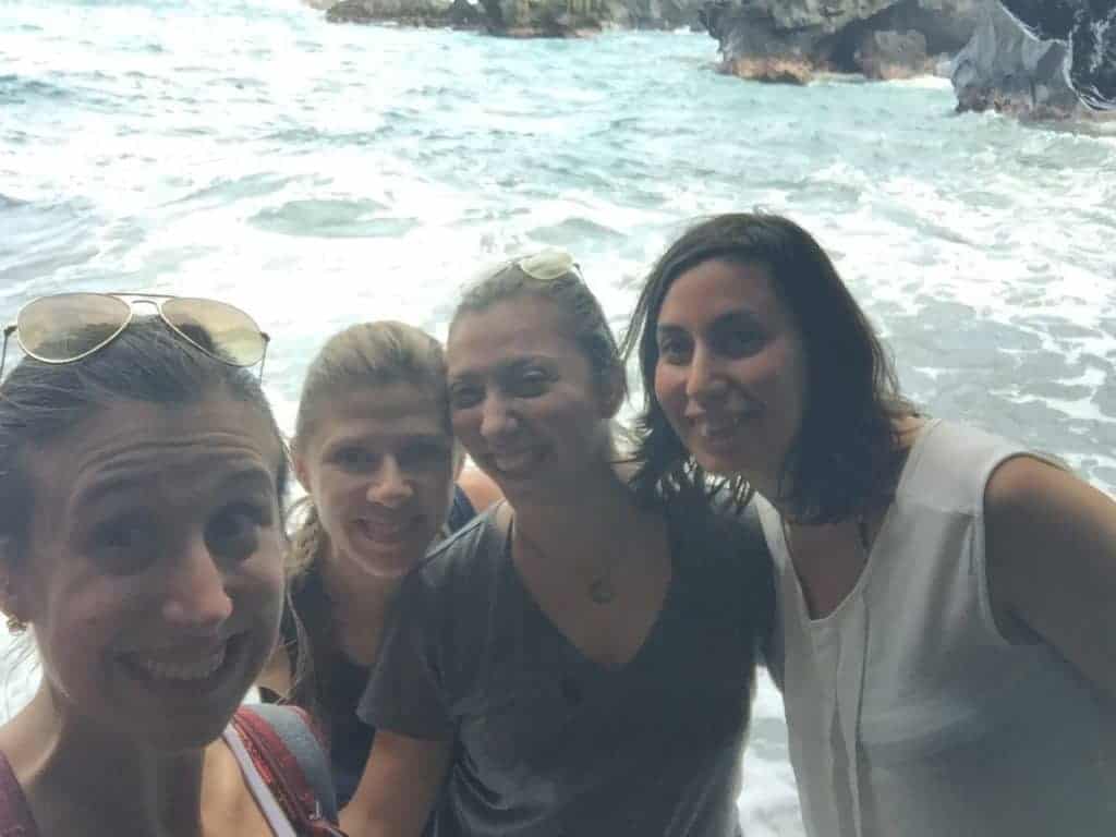 Four women stand close together and smile at the camera in front of a rocky shoreline with waves in the background. The lighting is bright and there are rocks and water behind them.