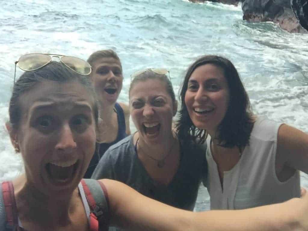 Four women stand close together near rocky ocean waves, smiling and making surprised faces at the camera. The background shows water splashing and dark rocks.
