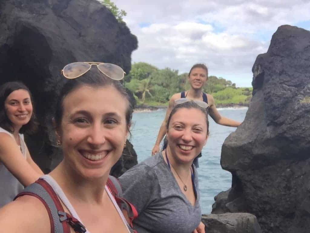 Four women stand on dark rocks by the water, smiling at the camera. Green foliage and a cloudy sky are visible in the background. The setting appears to be a natural outdoor area with rocky terrain.
