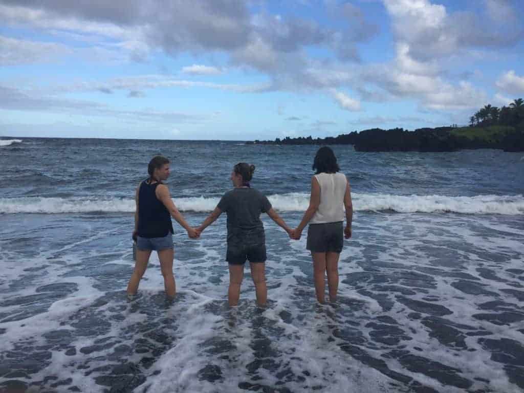 Three people stand in shallow ocean water, holding hands and facing away from the camera toward the horizon, with cloudy skies and dark, rocky shoreline in the background.