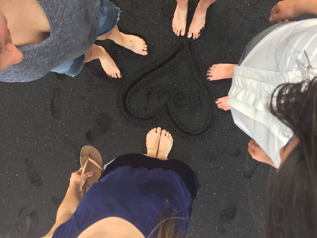 Five people stand barefoot in a circle on black sand, with their feet around a heart shape drawn in the sand that has the word "loved" inside it. One person holds a sandal. The photo is taken from above.