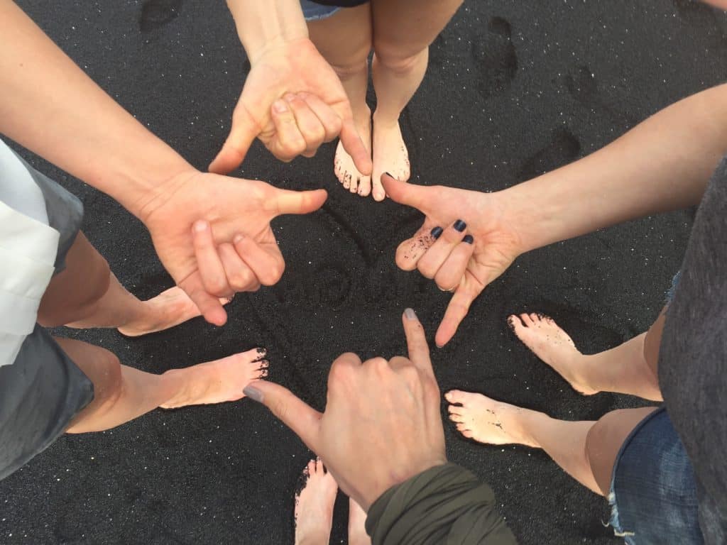 Five people standing barefoot on black sand, forming hand signs with their fingers pointing at each other in a circle. The sand and their legs are visible from above.