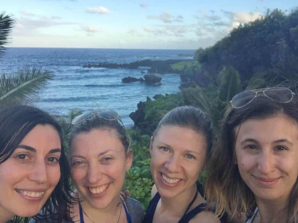 Four women pose for a selfie outdoors with the ocean, rocky coastline, and lush green vegetation in the background under a partly cloudy sky.