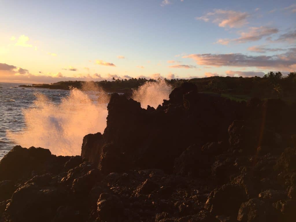 Waves crash against dark volcanic rocks at the shoreline during sunset, with sunlight illuminating the water spray and clouds visible in the sky above.