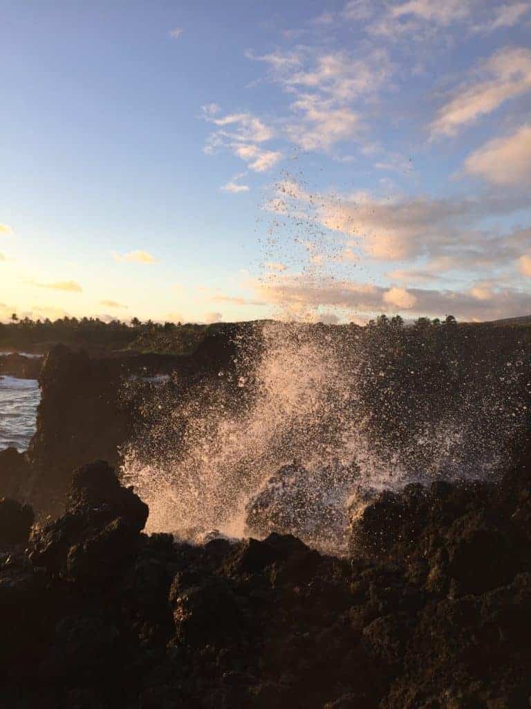 Waves crash against dark volcanic rocks on a coastline, sending water spray into the air under a mostly clear sky with scattered clouds at sunset. The landscape in the background is partially illuminated by the setting sun.