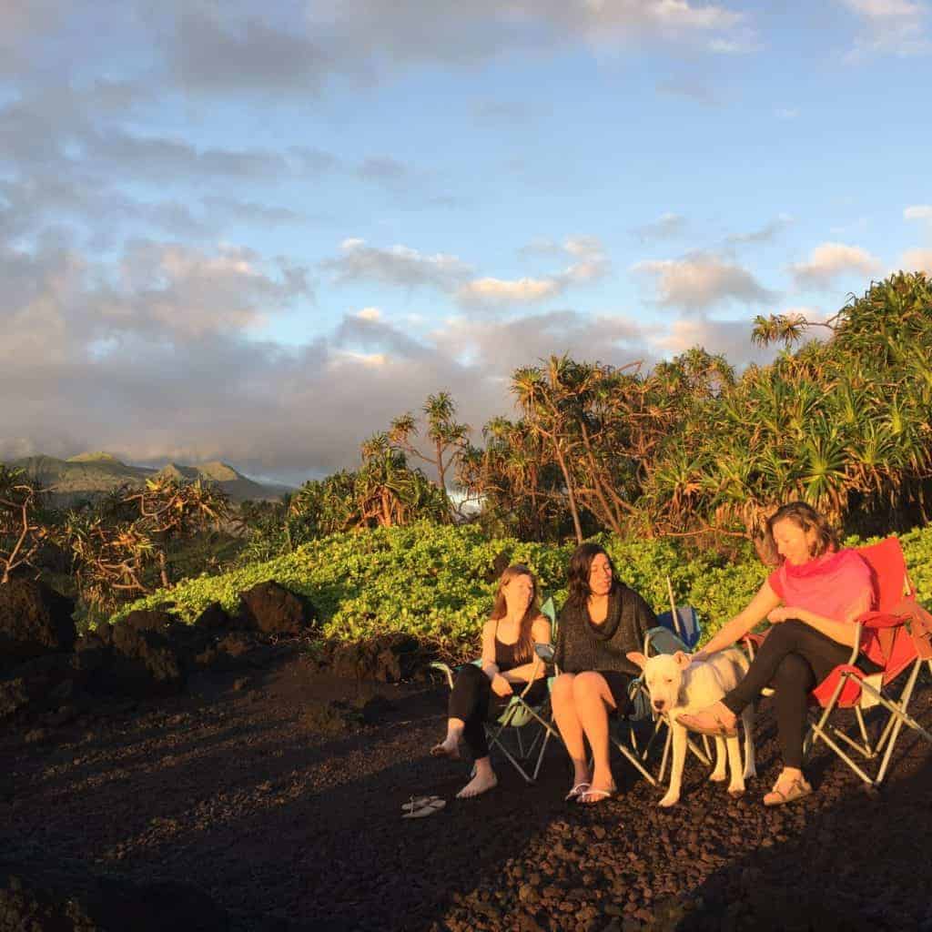 Three people sit in camping chairs on rocky ground outdoors, with one person holding a dog on a leash. There is greenery and mountains in the background under a cloudy sky.