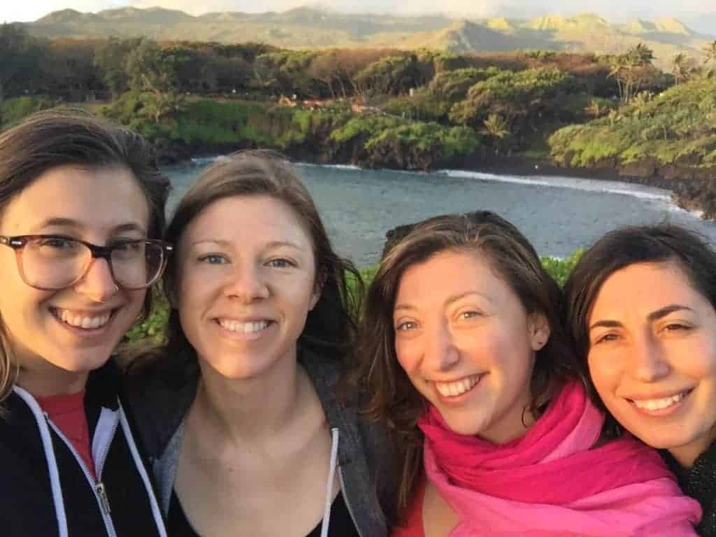 Four women smile at the camera in an outdoor setting with green hills, trees, and a body of water in the background under a partly cloudy sky.