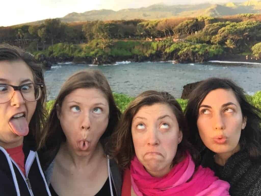 Four women stand outdoors in front of a body of water and green landscape, making exaggerated silly faces while looking upward or crossing their eyes. Trees and hills are visible in the background.