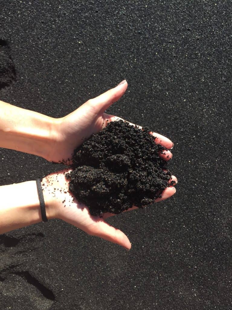 A person with light skin holds a mound of black sand in their hands over a surface covered with more black sand; a black hair tie is on their left wrist.