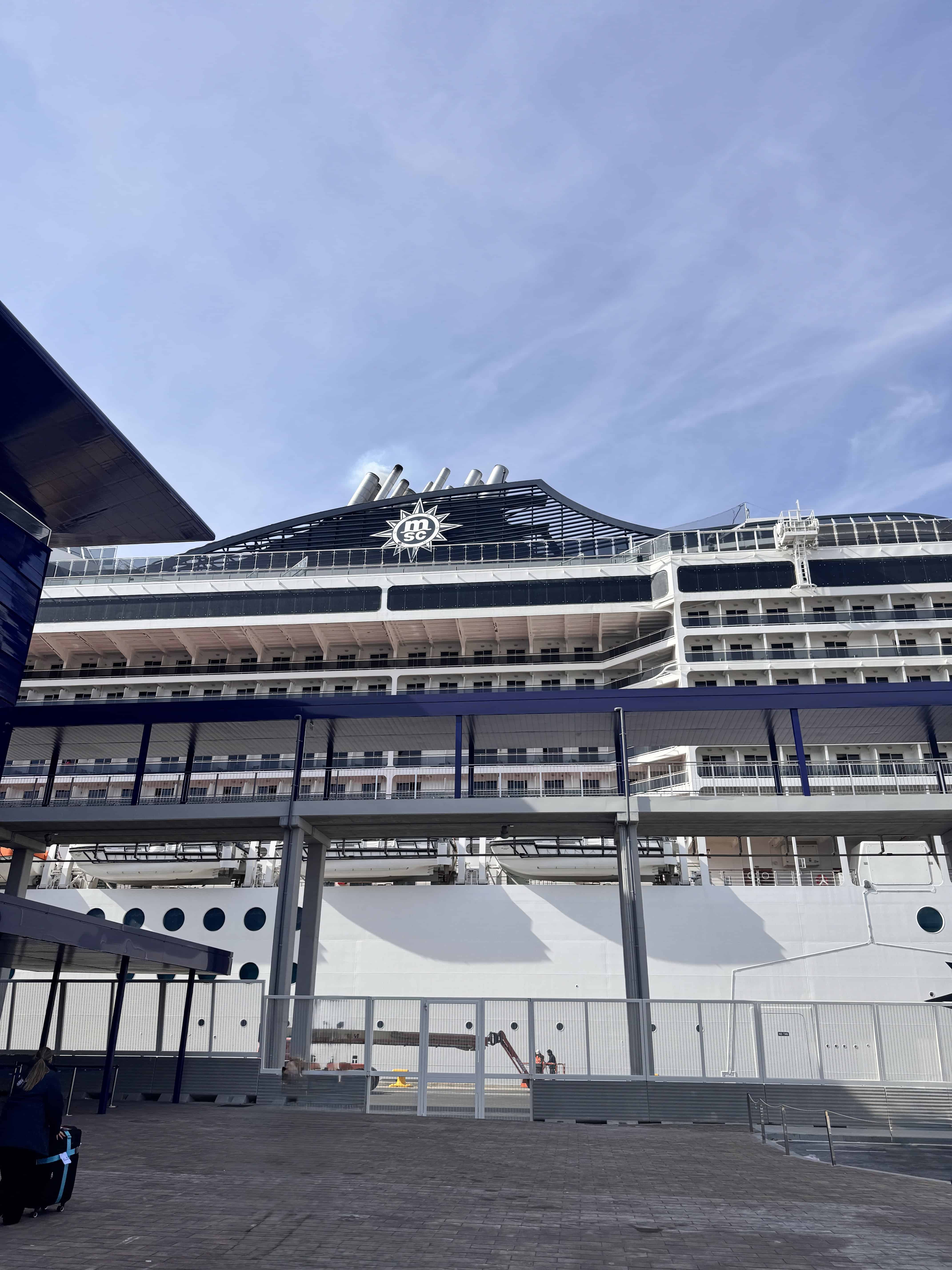 A large white cruise ship, MSC Fantasia, docked at a terminal with multiple decks visible and a covered walkway leading to the ship under a clear sky—perfect for your 2025 Cruise Review.