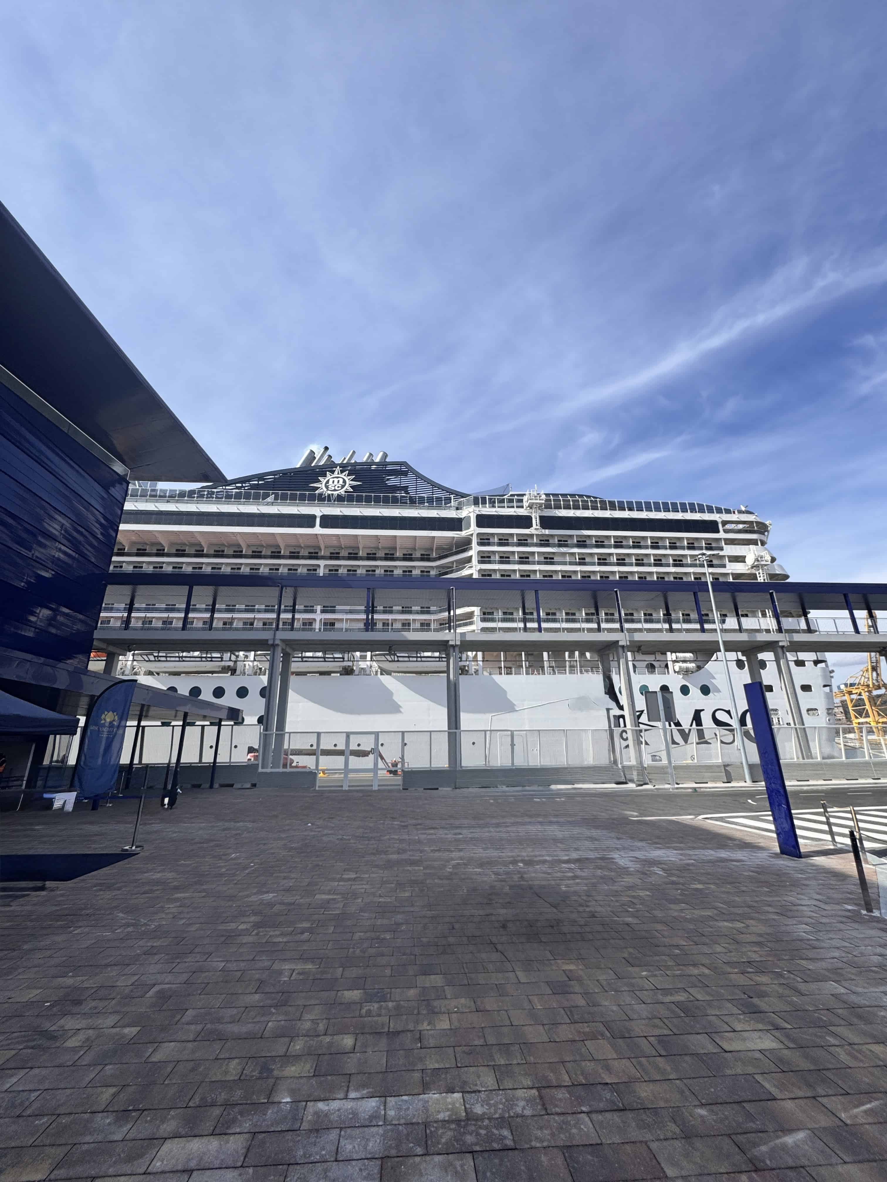 The MSC Fantasia cruise ship docked at a port, with buildings and a glass walkway in the foreground under a mostly blue sky—perfect for your 2025 Cruise Review.