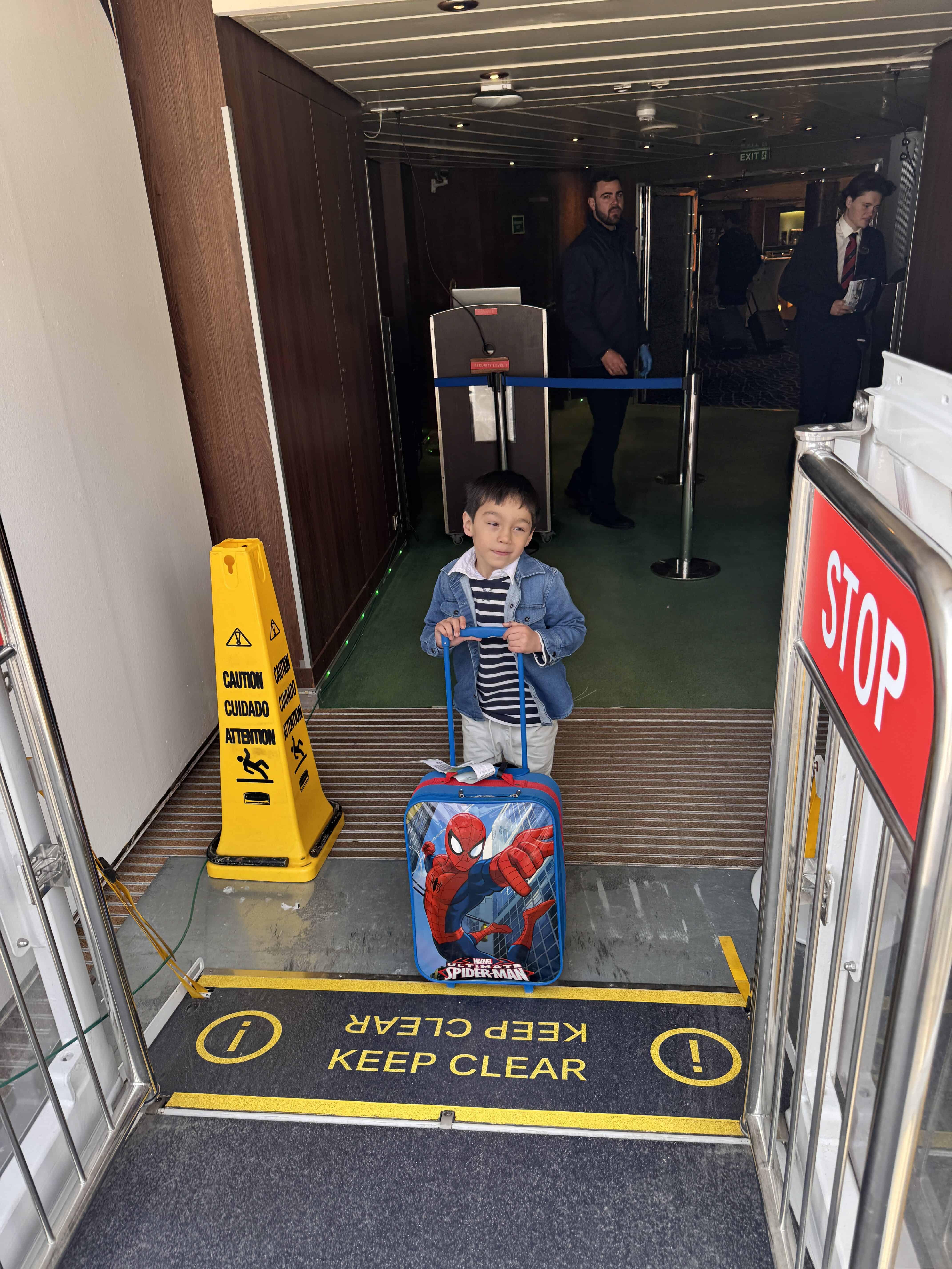 A young boy stands with a Spider-Man suitcase at an entrance marked "KEEP CLEAR," as two adults wait behind a barrier. Bright signs, including STOP and caution, hint at the excitement of a 2025 MSC Fantasia cruise review adventure ahead.