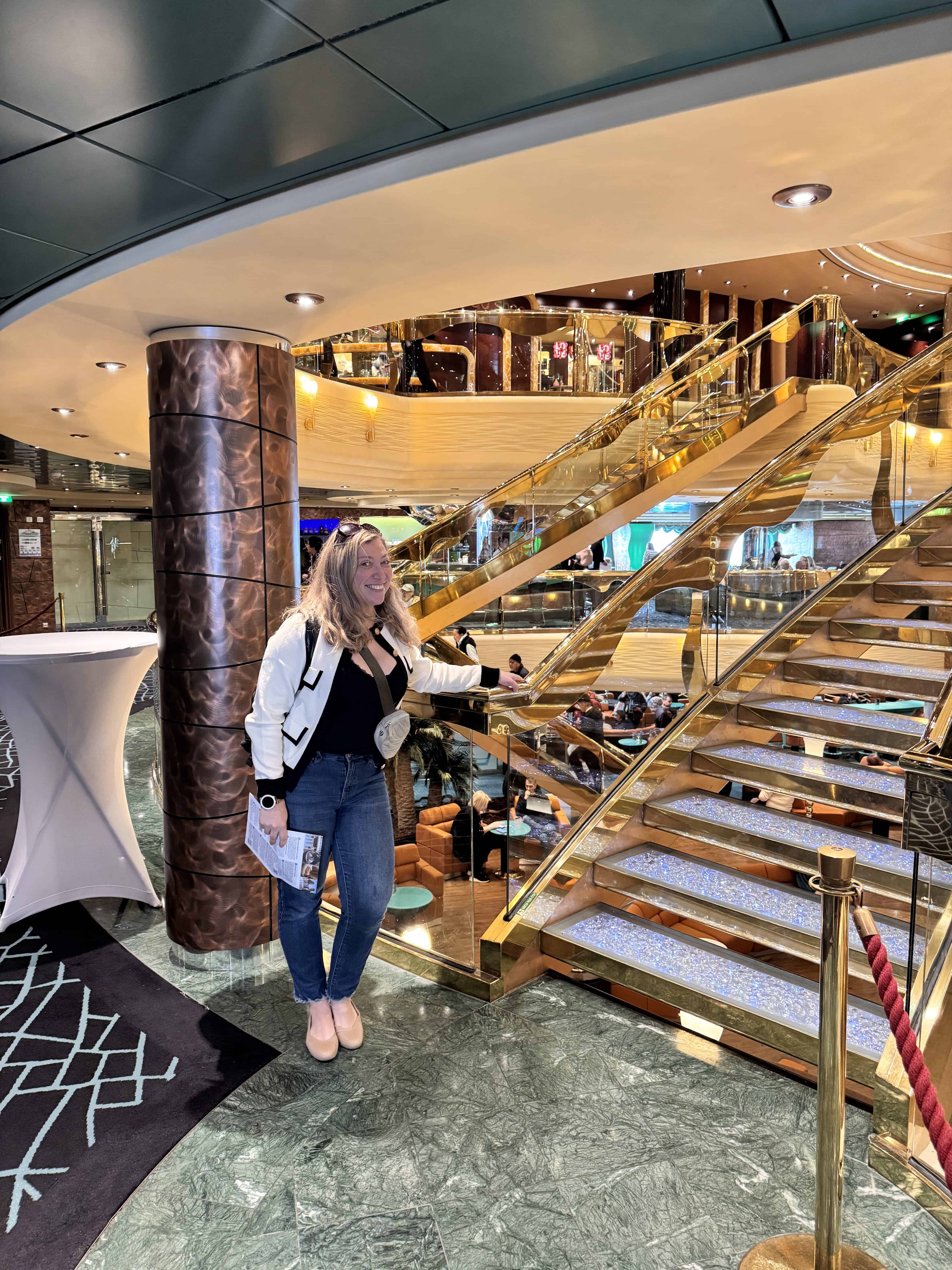 A woman with blonde hair stands next to a gold and glass staircase aboard the MSC Fantasia. Set against green marble floors and copper columns, she smiles while holding her phone—ready to capture her 2025 cruise review, dressed in jeans and a white jacket.