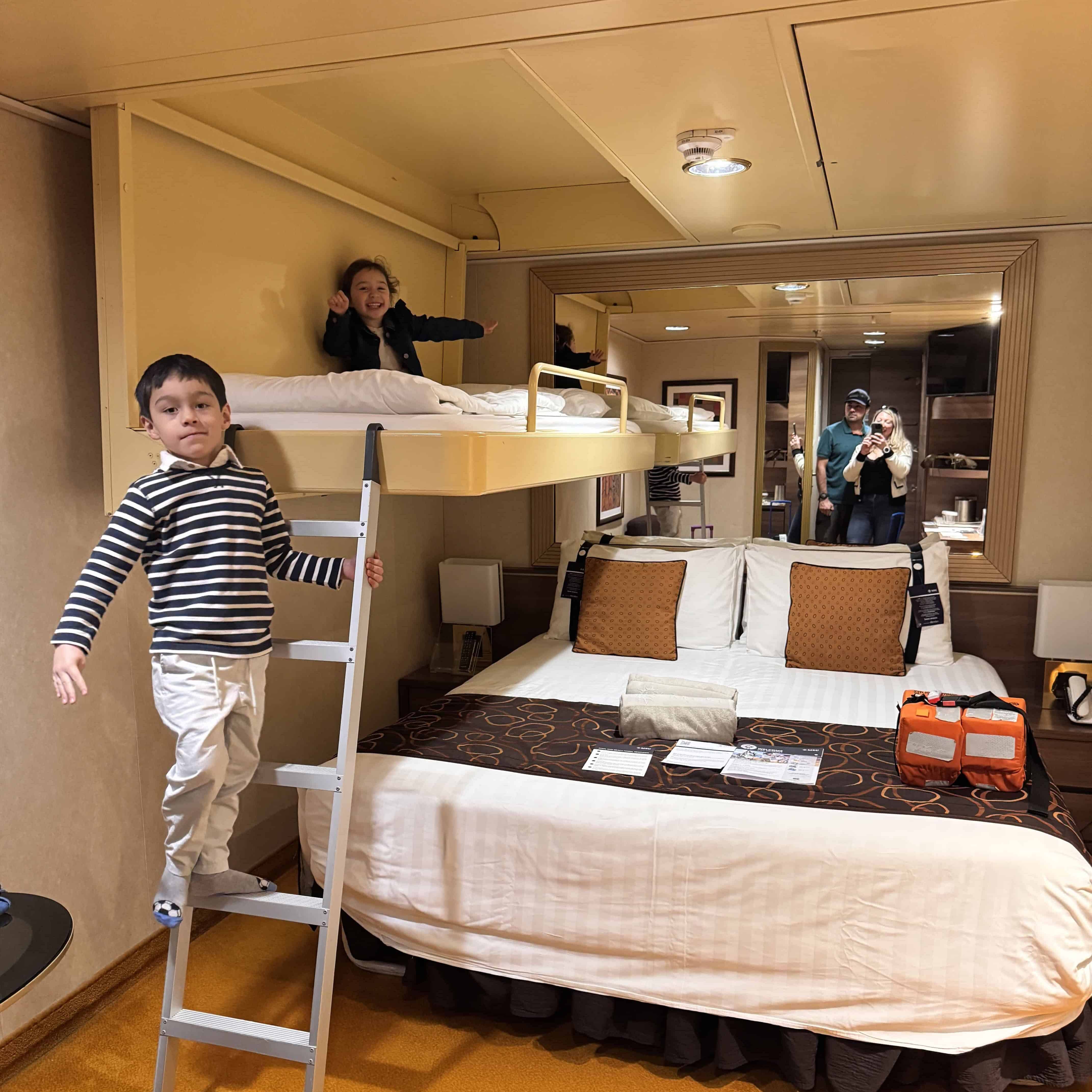 Two children play on a bunk bed above a double bed in an MSC Fantasia cruise ship cabin. One stands on the ladder while the other is on the top bunk, with two adults visible in the mirror’s reflection. Perfect for a 2025 Cruise Review!.