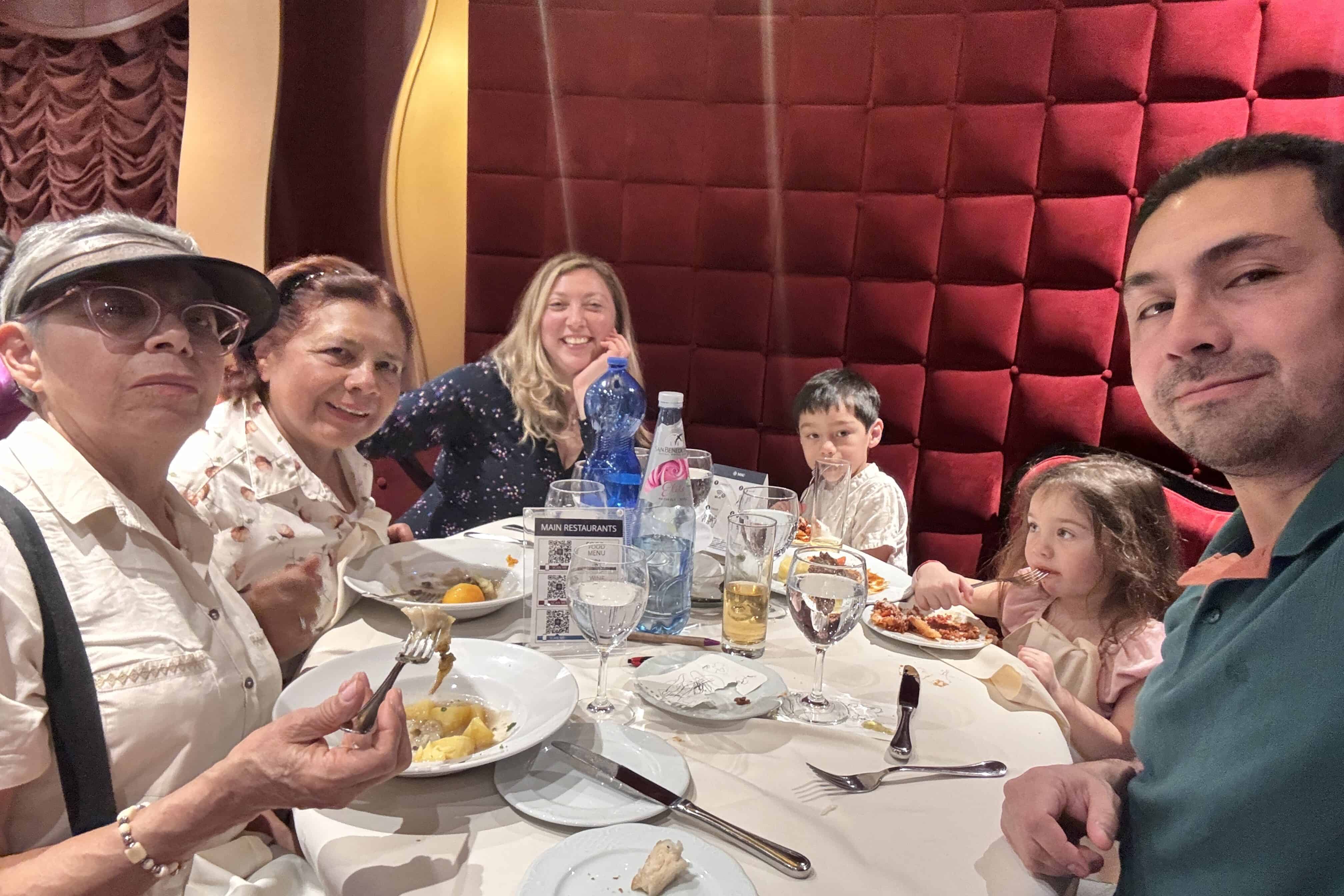 Five adults and two young children sit around a round dining table with plates of food and drinks in an MSC Fantasia restaurant with red padded walls, posing for a group photo and looking at the camera during their 2025 cruise review.