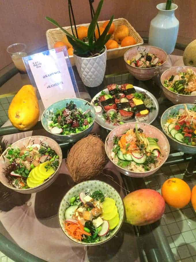A table displays several bowls of salads with assorted vegetables, a coconut, a yellow mango, an orange, a melon, a vase, a basket of oranges, and a potted plant. A glass of water and a sign are also present.