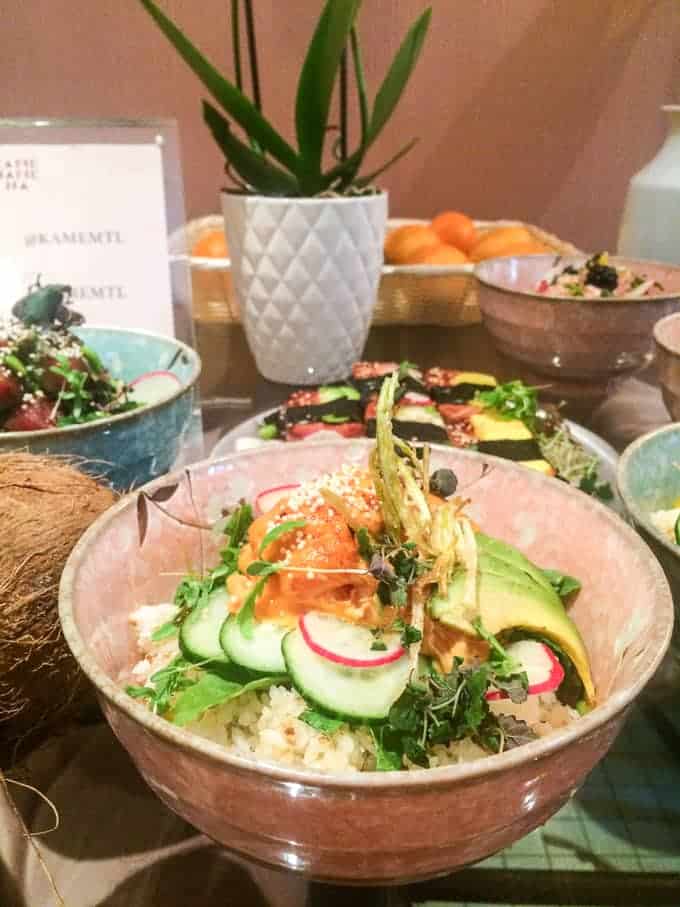 A bowl of rice topped with sliced cucumber, avocado, radish, greens, and sesame seeds is on a table with other dishes, a coconut, a plant in a white pot, and a basket of oranges in the background.