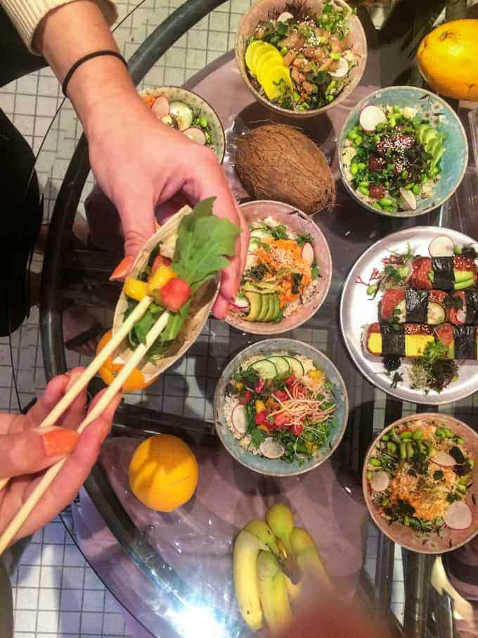 Overhead view of several bowls with colorful salads and sushi on a glass table. Two hands use chopsticks to pick up vegetables from a bowl. There are bananas, an orange, and a coconut also on the table.