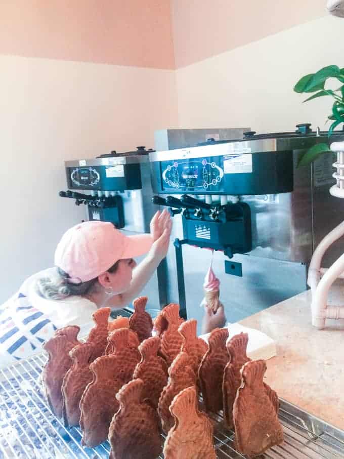 A person in a striped shirt and pink cap operates a soft-serve ice cream machine, holding a cone. In the foreground, several fish-shaped waffle cones are displayed on a cooling rack.