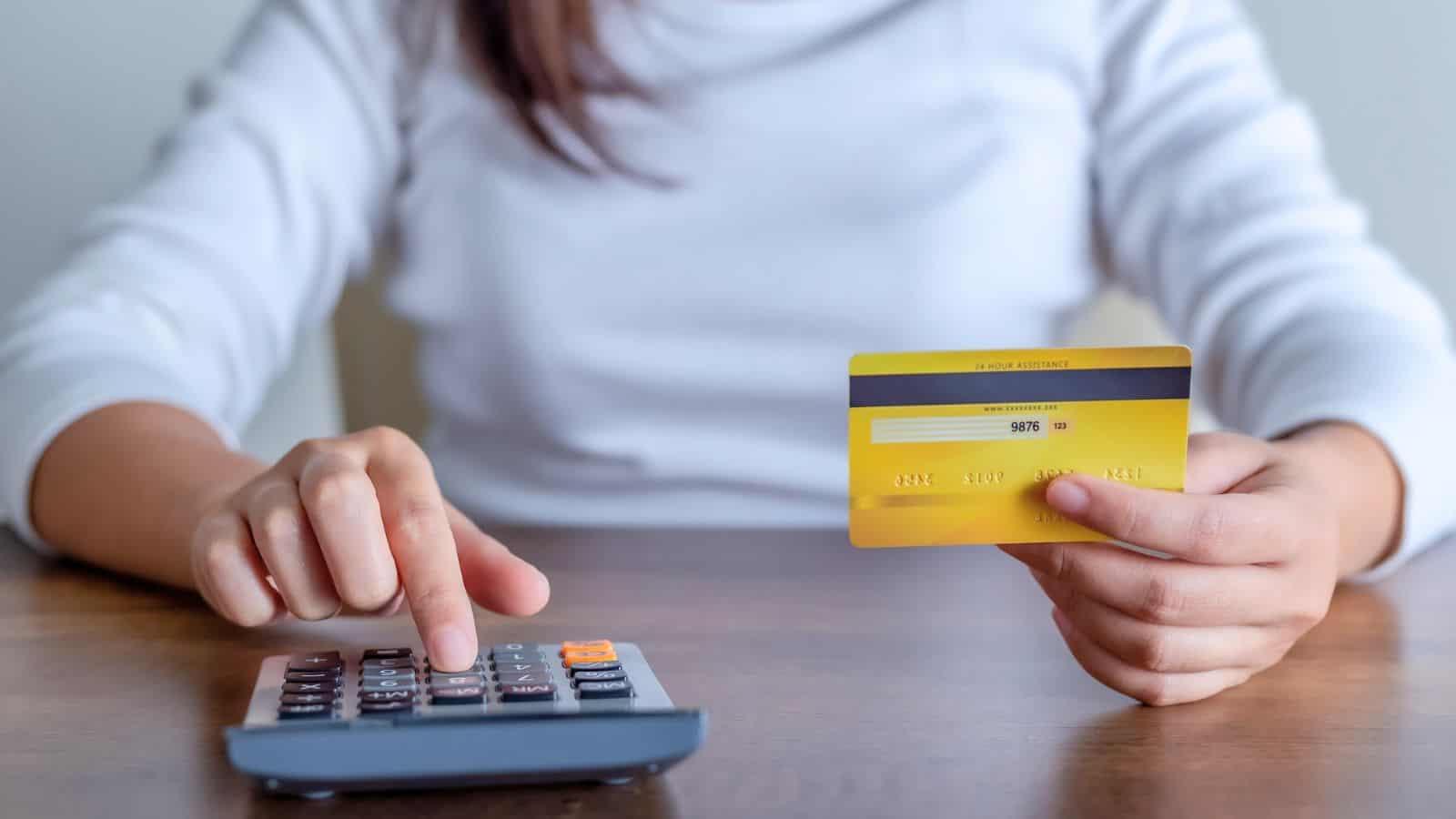 A person wearing a white long-sleeve shirt sits at a table, holding a yellow credit card in one hand and pressing buttons on a calculator with the other hand.