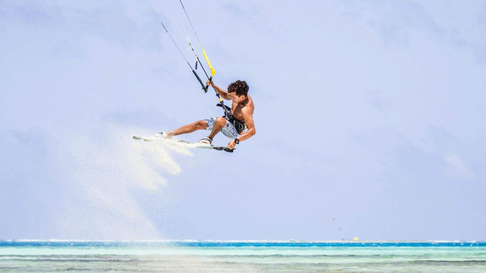 A person is kiteboarding over the ocean, high in the air and holding onto a control bar, with water spraying behind them. The sky is clear and the sea is calm with shades of blue and turquoise.