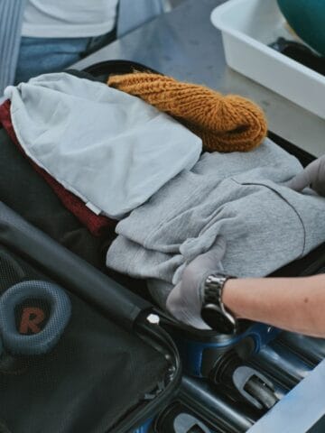 A security officer wearing gloves inspects an open suitcase at an airport security checkpoint. Inside the suitcase are folded clothes, a knitted hat, headphones, and a small bag. A neck pillow is in a tray nearby.