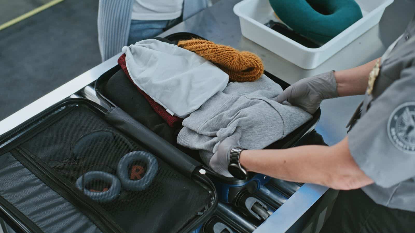 A security officer wearing gloves inspects an open suitcase at an airport security checkpoint. Inside the suitcase are folded clothes, a knitted hat, headphones, and a small bag. A neck pillow is in a tray nearby.