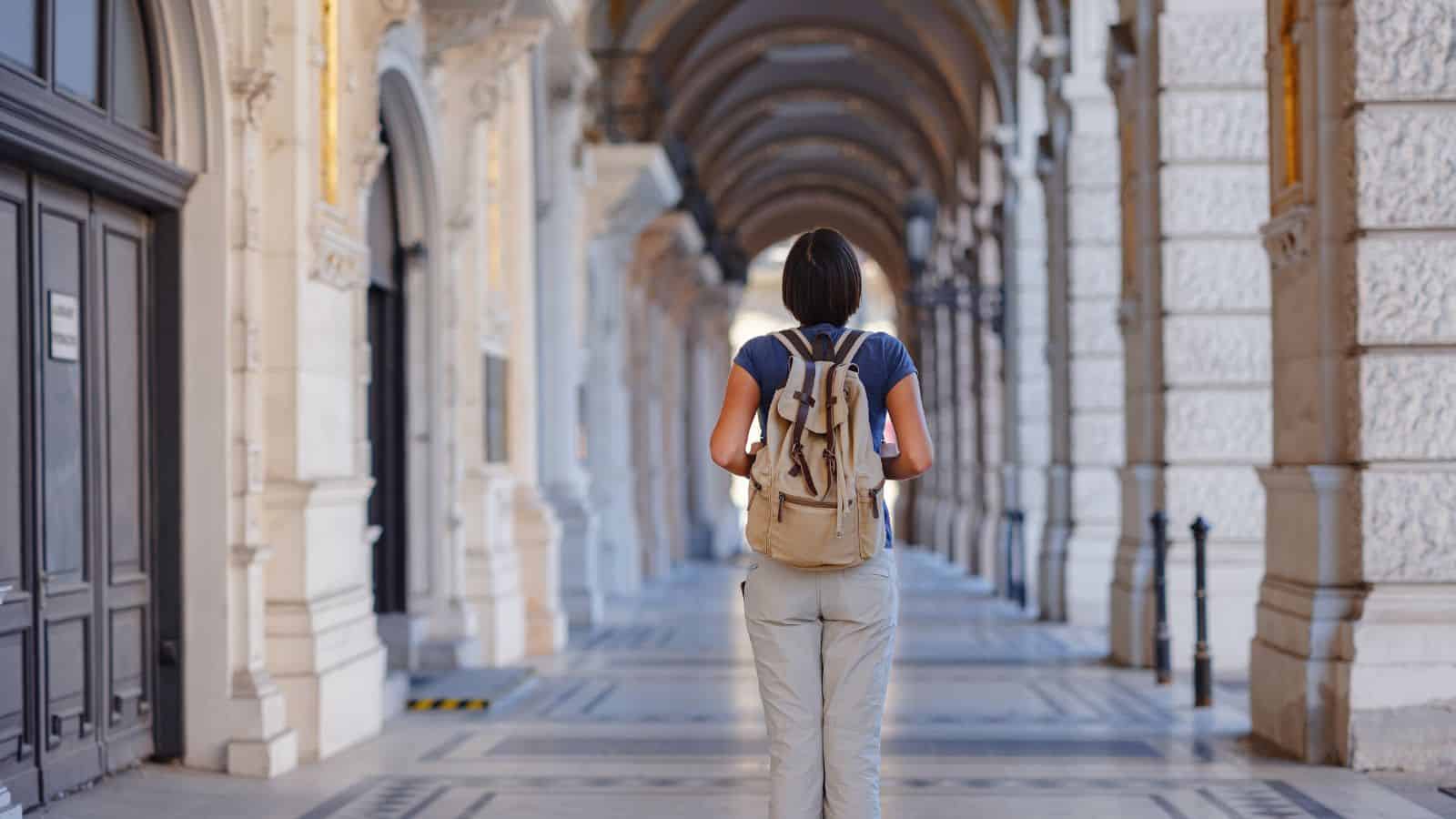 A person with short hair, wearing a blue shirt and beige pants, walks away from the camera under an ornate, arched corridor with columns and patterned flooring. The individual carries a light brown backpack.