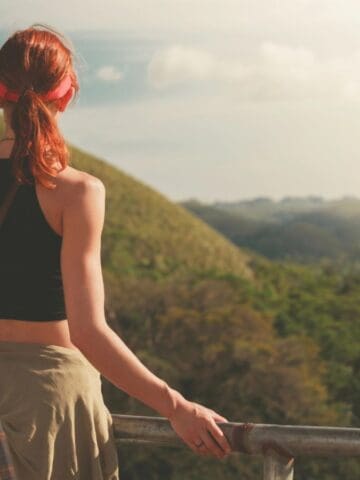 A person with red hair, wearing a black top and with a shirt tied around their waist, stands at a railing overlooking green hills and lush vegetation under a bright sky.