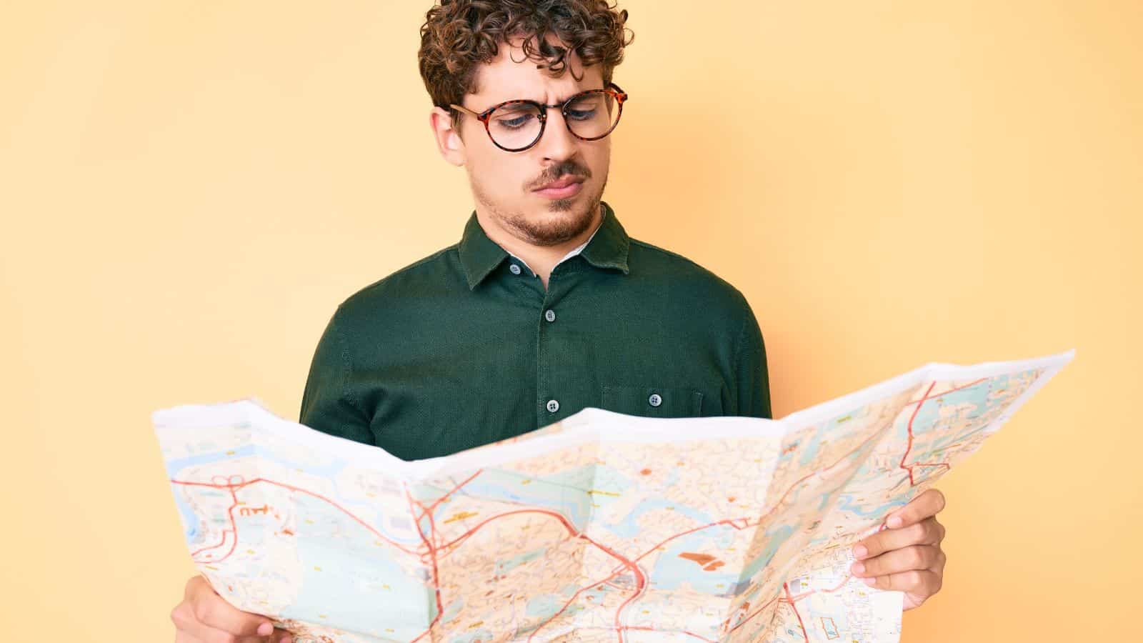A man with curly hair and glasses wearing a green shirt looks at a large folded map with a puzzled expression. The background is a solid light orange color.
