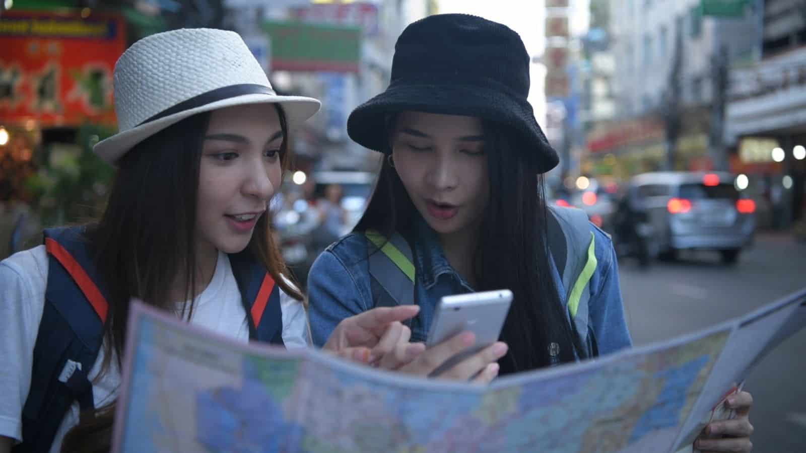 Two young women with backpacks stand on a busy city street, looking at a map and a smartphone. Both wear hats and casual clothes, appearing focused as they navigate or search for directions.