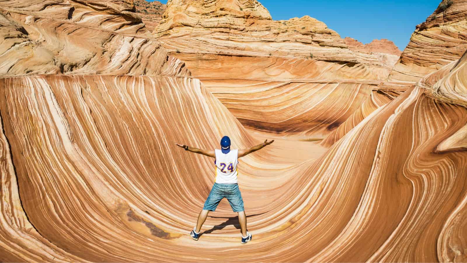 A person stands with arms outstretched facing layered, wavy sandstone formations at The Wave in Arizona under a clear blue sky. The rock has orange, tan, and white swirling patterns.