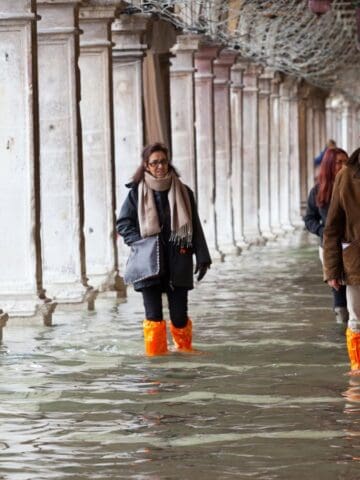 People walk through a flooded covered walkway, with water reaching their ankles. Most are wearing waterproof boots, including orange plastic covers. The columns and arches indicate an old, European architectural style.