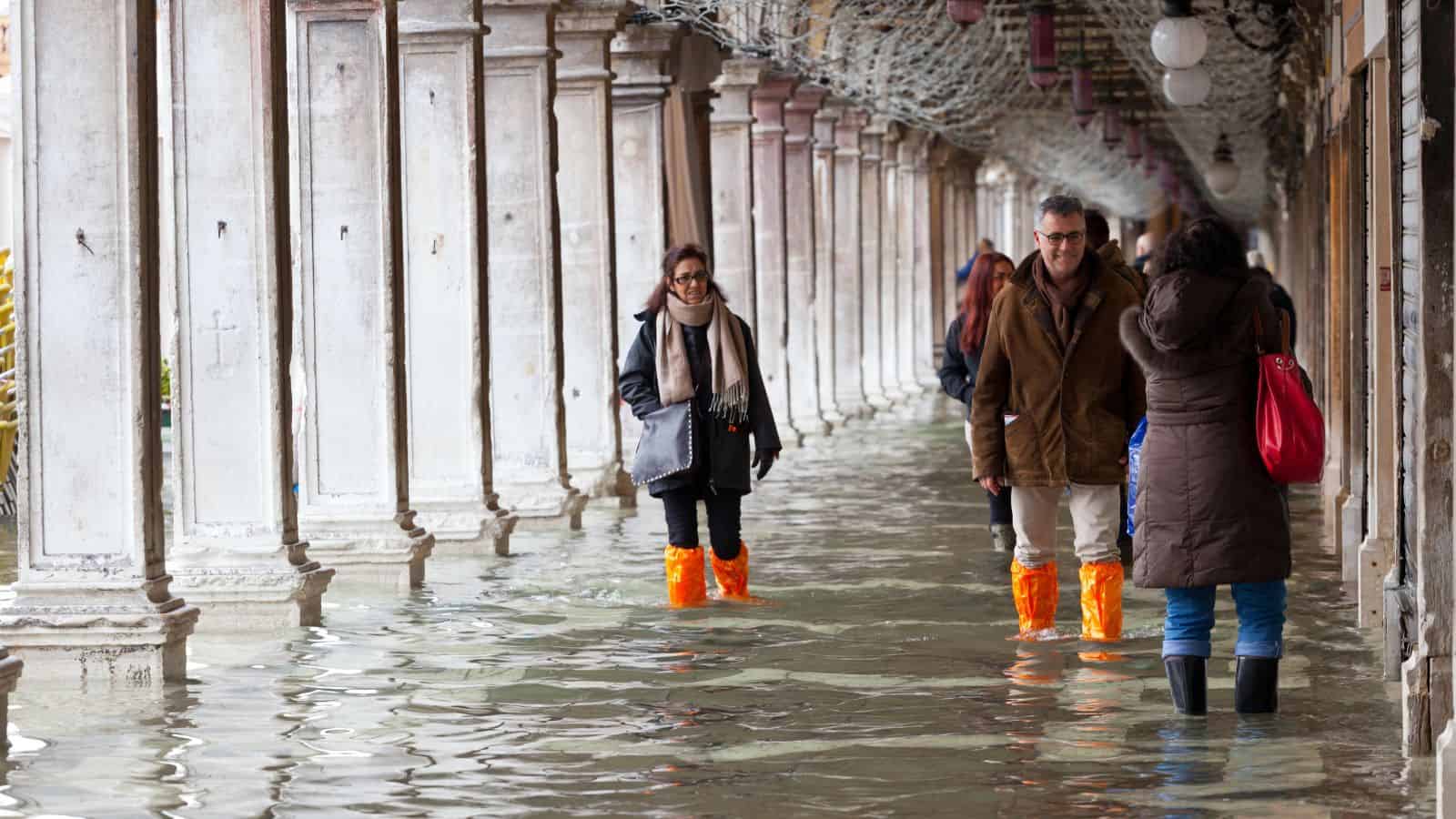 People walk through a flooded covered walkway, with water reaching their ankles. Most are wearing waterproof boots, including orange plastic covers. The columns and arches indicate an old, European architectural style.