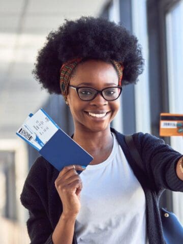 A person standing indoors holds up a boarding pass, passport, and credit card, smiling at the camera. Bright natural light comes through large windows in the background.