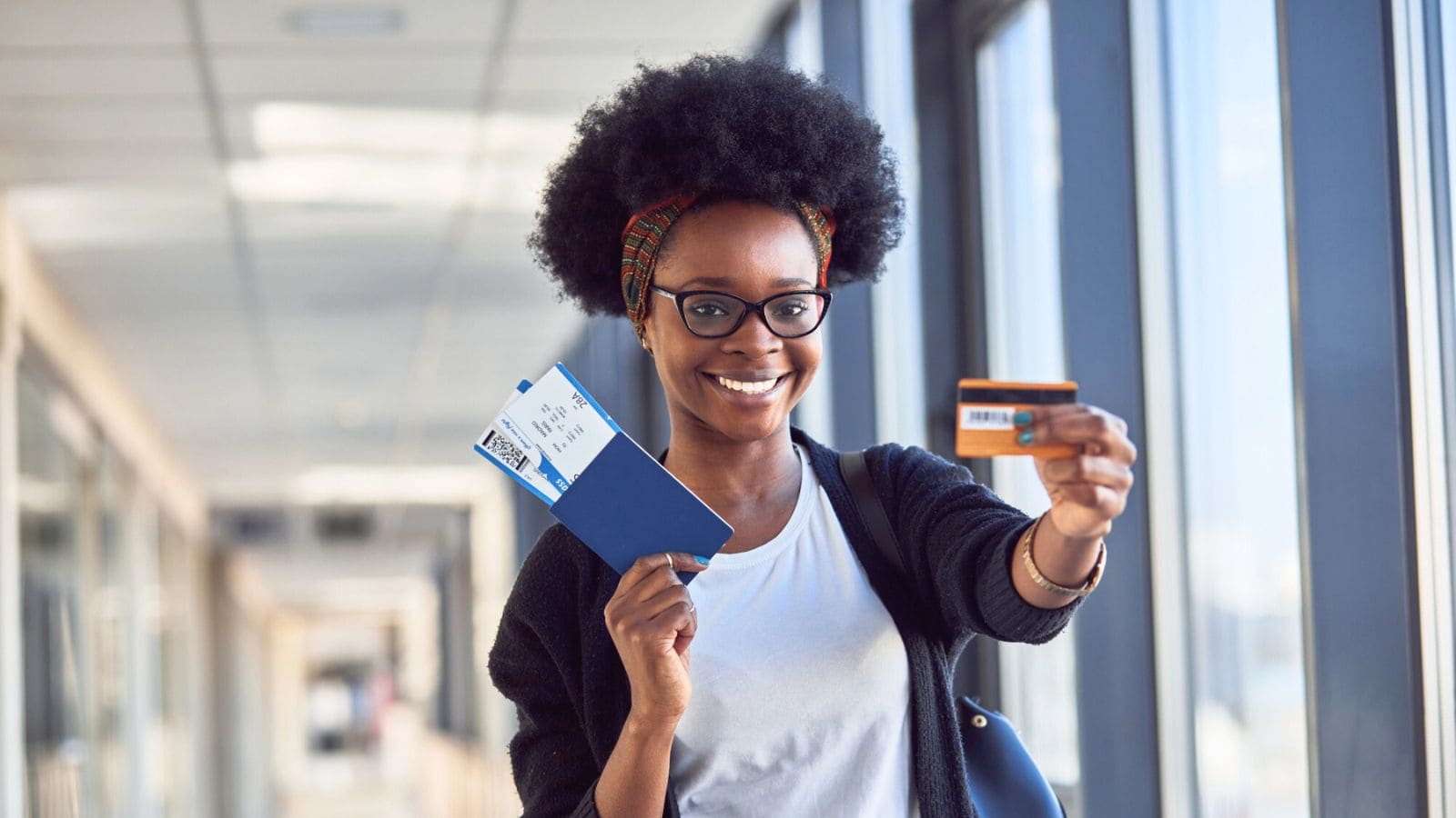 A person standing indoors holds up a boarding pass, passport, and credit card, smiling at the camera. Bright natural light comes through large windows in the background.