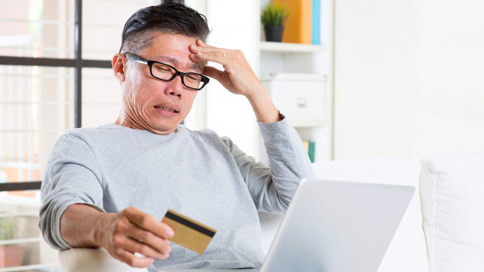 A person wearing glasses sits on a couch holding a credit card in one hand and touching their forehead with the other, looking at a laptop screen with a concerned expression.