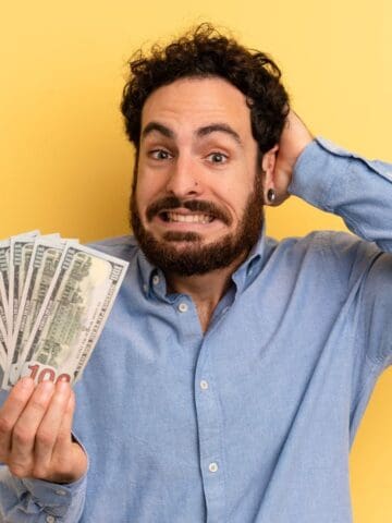 A man with curly hair and a beard, wearing a light blue shirt, holds a fan of U.S. dollar bills in one hand and touches the back of his head with the other, standing against a yellow background.